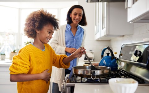 Pre-teen Mädchen steht am Herd in der Küche mit Spachtel und Pfanne, Zubereitung von Speisen mit ihrer Mutter, seitliche Ansich