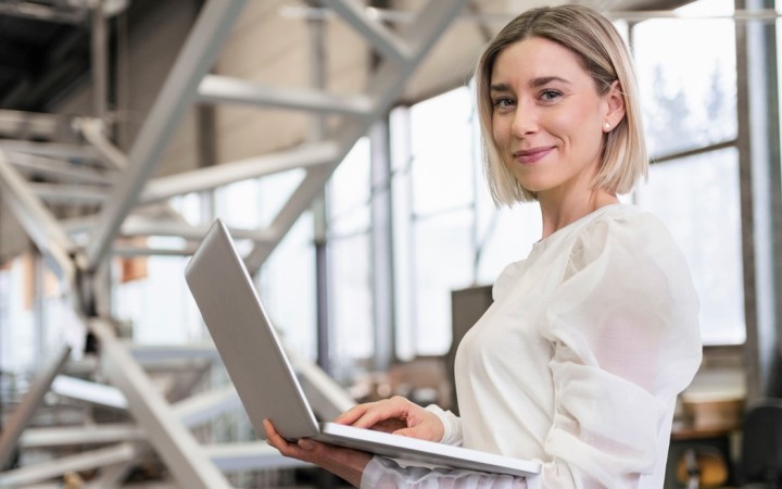 Blonde Frau mit kinnlangen Haaren und weißer Bluse steht mit einem Laptop in den Händen in einer Betriebshalle und lächelt in die Kamera.