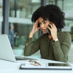 Frau mit Afro-Frisur stützt den Kopf in eine Hand, mit der anderen telefoniert sie, während sie vor einem Laptop sitzt und grübelnd darauf schaut