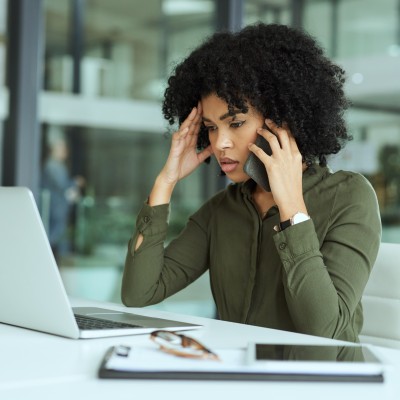 Frau mit Afro-Frisur stützt den Kopf in eine Hand, mit der anderen telefoniert sie, während sie vor einem Laptop sitzt und grübelnd darauf schaut