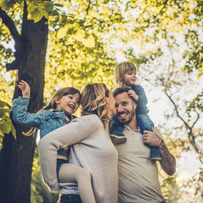 Familie mit zwei Kindern auf Rücken und Schultern geht lachend im  Wald bei Sonnenschein spazieren