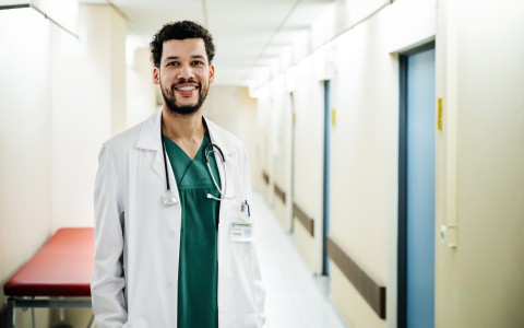 Ein junger Arzt steht in einem Krankenhausflur und lächelt in die Kamera
