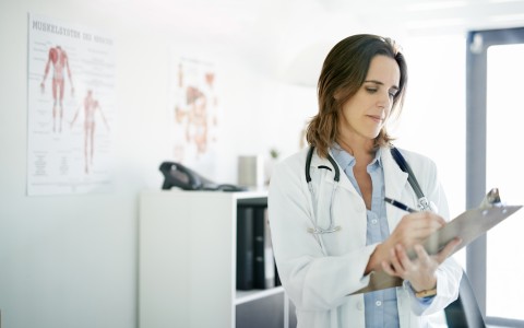 Ärztin steht in einem beratungszimmer und schreibt auf ein Klemmbrett
