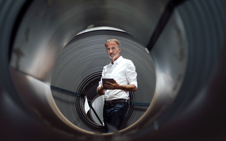 Mann mit weißem Hemd und grauem Bart steht mit einem Tablet in der Hand vor einer schwarzen Röhre