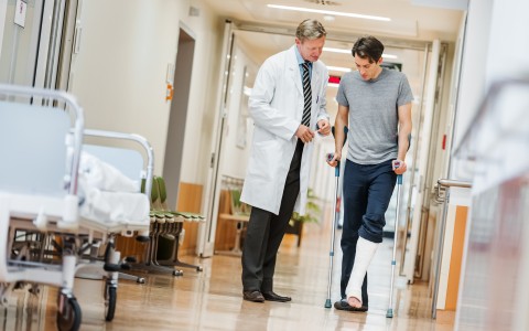 Ein Mann mit eingegipstem Fuß und Krücken steht in einem Krankenhauskorridor und redet mit einem Arzt