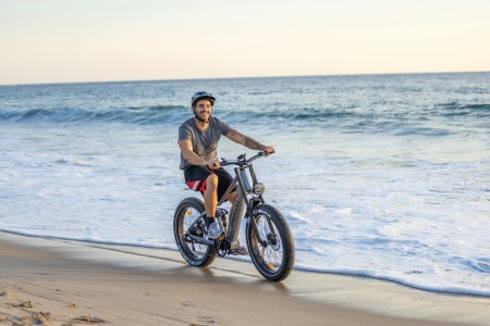 Junger Mann mit Helm fährt lachend auch einem E-Bike am Strand entlang