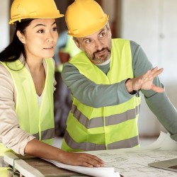 Frau und Mann in Schutzhelm und Warnweste besprechen einen Bauplan auf einer Baustelle